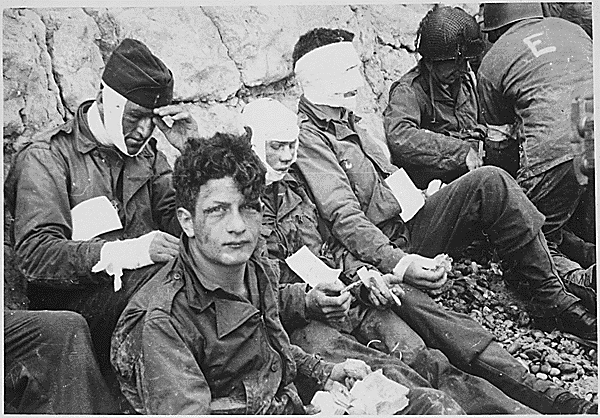 American assault troops of the 16th Infantry Regiment, injured while storming Omaha Beach, wait by the Chalk Cliffs for evacuation to a field hospital for further medical treatment. Collville-sur-Mer, Normandy, France., 06/06/1944. Credit: National Archives.