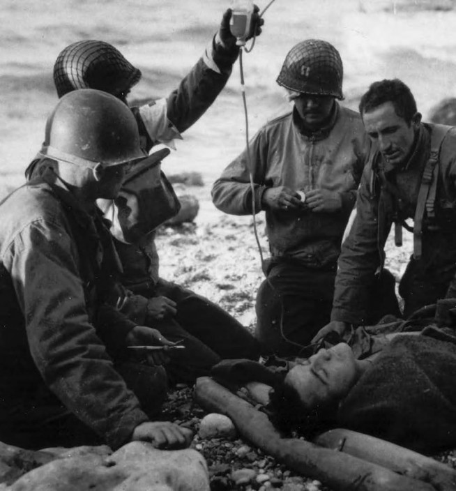 Army medics administering blood plasma to a survivor of a sunken landing craft on omaha Beach. D-Day casualties for the V Corps were in the neighborhood of 3,000 killed, wounded, and missing. The two assaulting regimental combat teams lost about 1,000 men each. The highest proportionate losses were taken by units that landed in the first few hours, including engineers, tank troops, and artillerymen. The D-Day casualties of V Corps were much higher than those suffered by VII Corps, where the assaulting seaborne division lost 197 men, including 60 lost at sea. Image and caption credit: Center of Military History. U.S. Army.