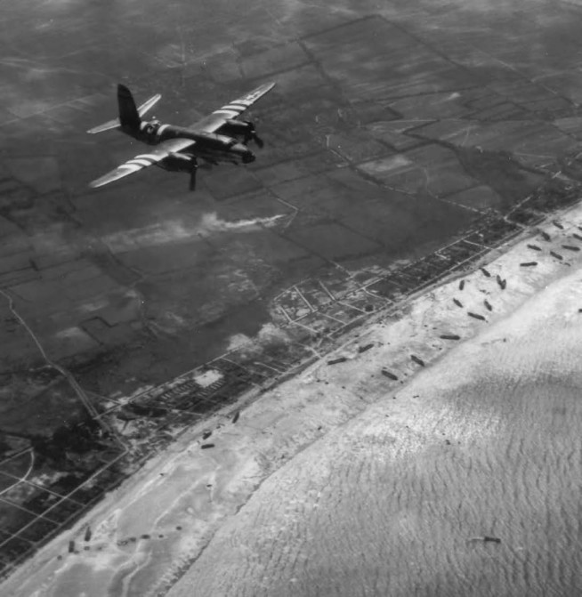 A Martin B-26 Medium Bomber flying over one of the invasion beaches, early on D-Day morning. All planes which supported the invasion operations, with the exception of the four-motored bombers, were painted with three white and two black stripes for identification purposes. At dawn on D Day the U. S. Air Forces took up the air attacks and in the half hour before the touchdown of the assault forces (from 0600 to 0630) 1,365 heavy bombers dropped 2,746 tons of high explosives on the shore defenses. This was followed by attacks by medium bombers, light bombers, and fighter bombers. During the 24 hours of 6 June Allied aircraft flew 13,000 sorties, and during the first 8 hours alone dropped 10,000 tons of bombs. Image and caption credit: Center of Military History. U.S. Army.