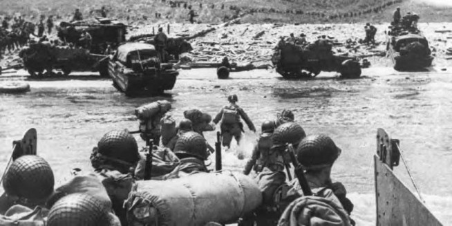 Troops leaving an LCVP to wade ashore. Half-tracks and 21/2-ton amphibian trucks can be seen on the beach, and in the background men marching in columns start southward toward the bluffs. On the shelf the enemy strung barbed wire and planted mines. Lanes had to be cleared through these obstacles before the infantry could advance. Beyond this strip containing obstacles, the enemy laid out firing positions to cover the tidal flat and the beach with direct fire, both plunging and grazing, from all types of weapons. The men landing were fired upon from these positions, which for the most part had escaped destruction during the prelanding bombardment. Image and caption credit: Center of Military History. U.S. Army.