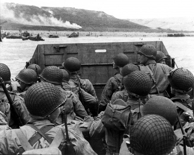 American assault troops in a landing craft huddle behind the protective front of the craft as it nears a beachhead, on the Northern Coast of France. Smoke in the background is Naval gunfire supporting the land. Image and credit: U.S. Army Center of Military History.