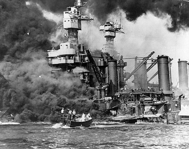 Sailors in a motor launch rescue a survivor from the water alongside the sunken USS West Virginia (BB-48) during or shortly after the Japanese air raid on Pearl Harbor. USS Tennessee (BB-43) is inboard of the sunken battleship. Note extensive distortion of West Virginia's lower midships superstructure, caused by torpedoes that exploded below that location. Also note 5"/25 gun, still partially covered with canvas, boat crane swung outboard and empty boat cradles near the smokestacks, and base of radar antenna atop West Virginia's foremast. Official U.S. Navy Photograph, National Archives Collection. Caption: Naval History & Heritage Command.