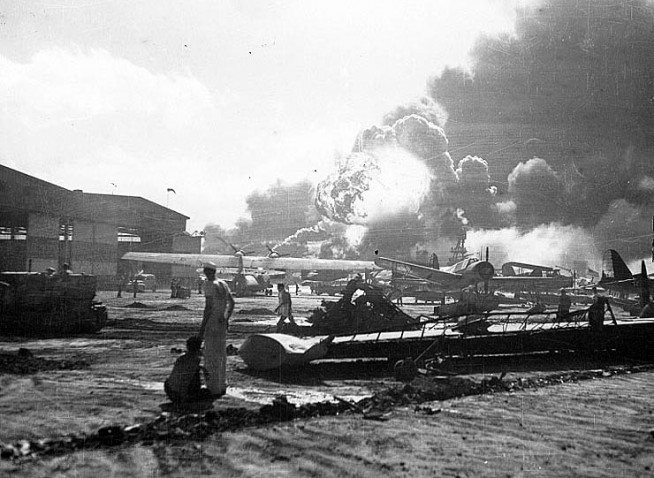 Sailors stand amid wrecked planes at the Ford Island seaplane base, watching as USS Shaw (DD-373) explodes in the center background, 7 December 1941. USS Nevada (BB-36) is also visible in the middle background, with her bow headed toward the left. Planes present include PBY, OS2U and SOC types. Wrecked wing in the foreground is from a PBY. Official U.S. Navy Photograph, National Archives Collection. Caption: Naval History & Heritage Command.