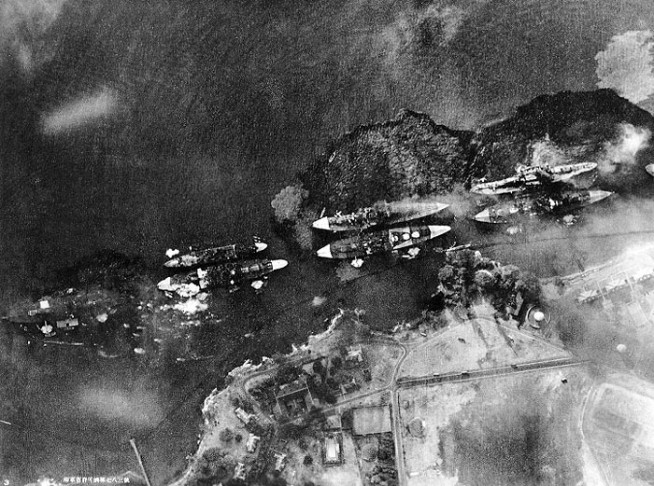 Vertical aerial view of "Battleship Row", beside Ford Island, during the early part of the horizontal bombing attack on the ships moored there. Photographed from a Japanese aircraft. Ships seen are (from left to right): USS Nevada ; USS Arizona with USS Vestal moored outboard; USS Tennessee with USS West Virginia moored outboard; USS Maryland with USS Oklahoma moored outboard; and USS Neosho, only partially visible at the extreme right. A bomb has just hit Arizona near the stern, but she has not yet received the bomb that detonated her forward magazines. West Virginia and Oklahoma are gushing oil from their many torpedo hits and are listing to port. Oklahoma's port deck edge is already under water. Nevada has also been torpedoed. Japanese inscription in lower left states that the photograph has been officially released by the Navy Ministry. Photo and caption: National History & Heritage Command.