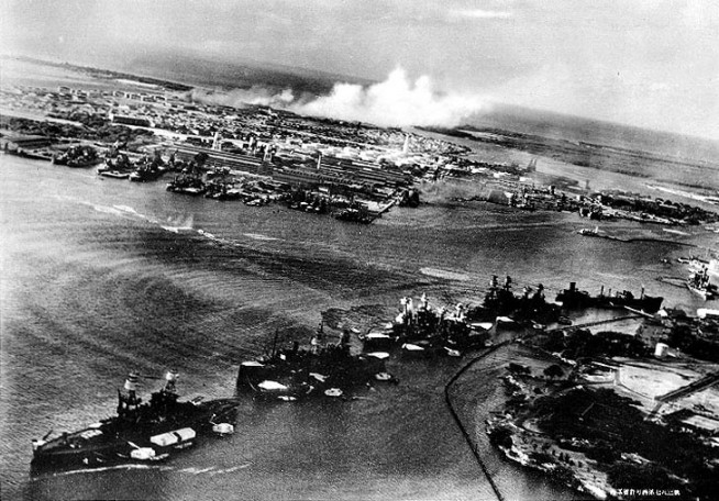 Torpedo planes attack "Battleship Row" at about 0800 on 7 December, seen from a Japanese aircraft. Ships are, from lower left to right: Nevada (BB-36) with flag raised at stern; Arizona (BB-39) with Vestal (AR-4) outboard; Tennessee (BB-43) with West Virginia (BB-48) outboard; Maryland (BB-46) with Oklahoma (BB-37) outboard; Neosho (AO-23) and California (BB-44). West Virginia, Oklahoma and California have been torpedoed, as marked by ripples and spreading oil, and the first two are listing to port. Torpedo drop splashes and running tracks are visible at left and center. White smoke in the distance is from Hickam Field. Grey smoke in the center middle distance is from the torpedoed USS Helena (CL-50), at the Navy Yard's 1010 dock. Japanese writing in lower right states that the image was reproduced by authorization of the Navy Ministry. Photo and caption: Naval Heritage & History Command.