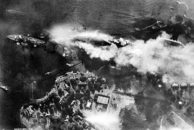 Vertical aerial view of "Battleship Row", beside Ford Island, soon after USS Arizona was hit by bombs and her forward magazines exploded. Photographed from a Japanese aircraft. Ships seen are (from left to right): USS Nevada; USS Arizona (burning intensely) with USS Vestal moored outboard; USS Tennessee with USS West Virginia moored outboard; and USS Maryland with USS Oklahoma capsized alongside. Smoke from bomb hits on Vestal and West Virginia is also visible. Japanese inscription in lower left states that the photograph has been reproduced under Navy Ministry authorization. Photo and caption: National History & Heritage Command.