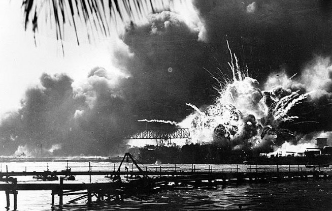 The forward magazine of USS Shaw (DD-373) explodes during the second Japanese attack wave. To the left of the explosion, Shaw's stern is visible, at the end of floating drydock YFD-2. At right is the bow of USS Nevada (BB-36), with a tug alongside fighting fires. Photographed from Ford Island, with a dredging line in the foreground. Photo and caption: Naval History & Heritage Command.