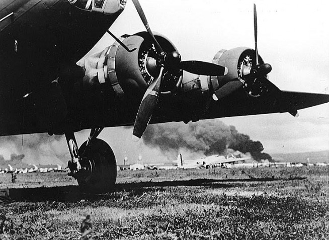 A U.S. Army B-17E at Hickam Air Field, after landing safely during the Japanese air raid. In the background is a B-17C (or B-17D). Smoke from burning ships at Pearl Harbor is visible in the distance. The B-17E is probably that piloted by First Lieutenant Karl T. Barthelmess. Photographer may be Staff Sergeant Lee Embree. Photograph from Army Signal Corps Collection, National Archives. Caption: Naval History & Heritage Command.