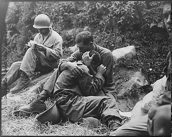 A grief stricken American infantryman whose buddy has been killed in action is comforted by another soldier. In the background a corpsman methodically fills out casualty tags, Haktong-ni area, Korea.: 08/28/1950. Credit: National Archives.