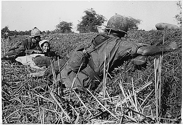 A soldier of the 1st Infantry Division motions to a woman refugee to keep her children's heads down during a fight with Viet Cong who had attempted to ambush the unit during a move through an area criss-crossed with bamboo hedgerows., 01/16/1966. Credit: National Archives.