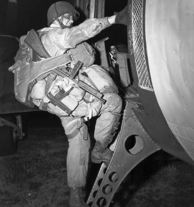 Fully Equipped paratrooper armed with a Thompson submachine gun M1, climbing into a transport plane to go to France as the invasion of Normandy gets under way. At approximately 0200, 6 June 1944, men of two U. S. airborne divisions, as well as elements of a British airborne division, were dropped in vital areas to the rear of German coastal defenses guarding the Normandy beaches from Cherbourg to Caen. By dawn 1,136 heavy bombers of the RAF Bomber Command had dropped 5,853 tons of bombs on selected coastal batteries lining the Bay of the Seine between Cherbourg and Le Havre. Image and caption credit: Center of Military History. U.S. Army.