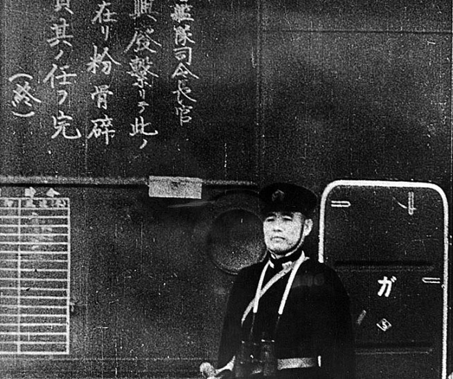 The Commanding Officer of the Japanese aircraft carrier Shokaku watches as planes take off to attack Pearl Harbor, during the morning of 7 December 1941. The Kanji inscription at left is an exhortation to pilots to do their duty. Official U.S. Navy Photograph, National Archives Collection. Caption: Naval History and Heritage Command.