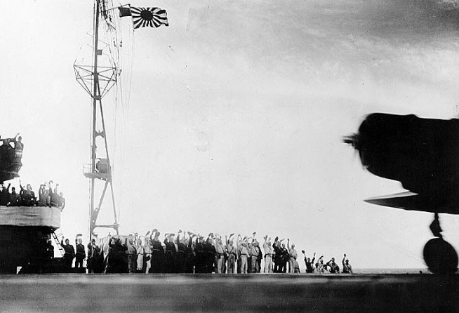 A Japanese Navy Type 97 Carrier Attack Plane ("Kate") takes off from a carrier as the second wave attack is launched. Ship's crewmen are cheering "Banzai" This ship is either Zuikaku or Shokaku. Note light tripod mast at the rear of the carrier's island, with Japanese naval ensign. Photo & Caption: Naval History & Heritage Command.