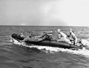 Clad in rubber exposure suit, a Marine scout swimmer dives from an inflatable boat more than five hundred yards offshore. The exposure suit is worn in cold climates where water temperatures hamper swimmers. Working in teams of two, the scout swimmers examine the beach for obstacles and enemy patrols. If the area is clear, they signal the remainder of the reconnaissance patrol to come ashore. Caption credit: MARSOC by Fred Pushies. Image credit: USMC HISTORY DIVISION, QUANTICO.