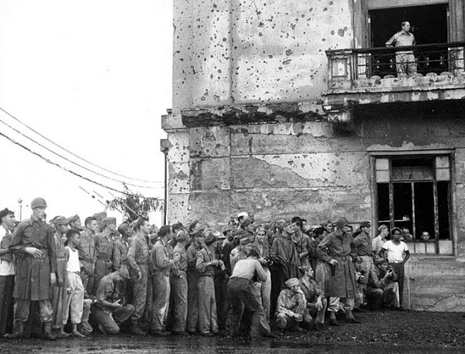 General of the Army Douglas MacArthur (top right) watches from a balcony above a crowd of soldier spectators as the sixteen-man Japanese delegation arrives at City Hall, Manila, to make surrender arrangements. Photo is dated 20 August 1945. Credit: Naval Historical Center.
